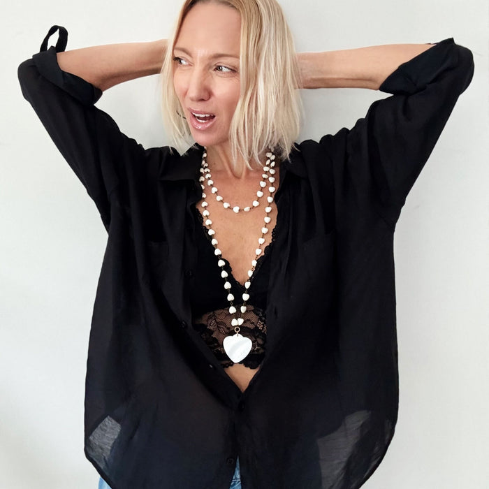 A smiling person with blonde hair wears a black shirt and layered necklaces, including a delicate layering chain adorned with howlite heart beads, against a plain background.