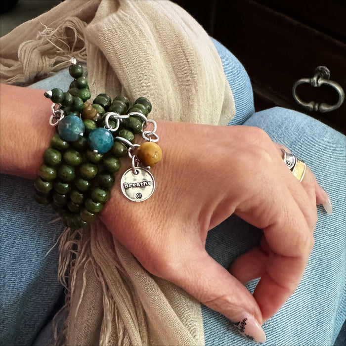A person's wrist wearing a boho-style wrap bracelet with green wooden beads and silver pendant, accompanied by a magnetic clasp.