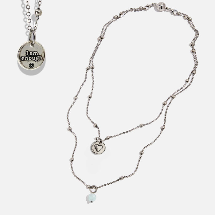 Two dainty necklaces with pendants, one with a silver finish and the other with a gold finish. Both have a chain link design and feature a circular pendant with the phrase 'I am enough' and a heart symbol respectively.