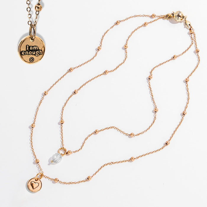 Two dainty necklaces with pendants, one with a silver finish and the other with a gold finish. Both have a chain link design and feature a circular pendant with the phrase 'I am enough' and a heart symbol respectively.