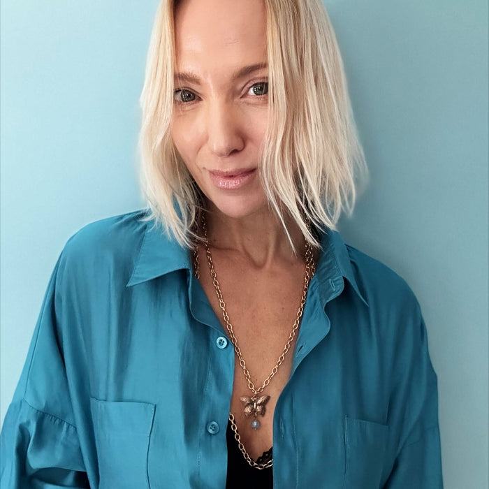Woman in teal shirt with tousled blonde bob, wearing a butterfly pendant and large ring, against a blue background.