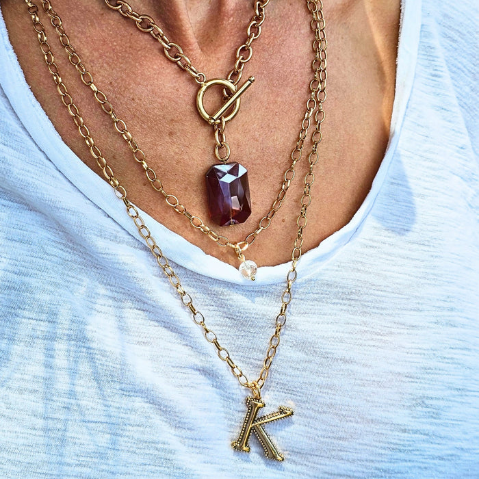 Gold necklace with a red gemstone pendant and 'K' pendant on a light blue top.