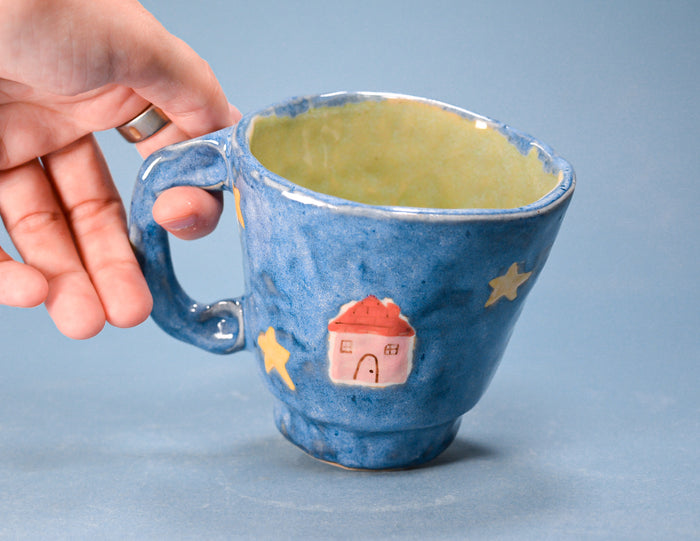 A hand holding a blue ceramic mug with a yellow-green glazed interior, decorated with yellow stars and a small pink house with a red roof.