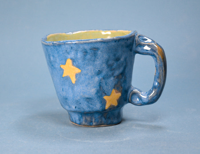 A handmade ceramic mug with a textured blue glaze, decorated with two yellow stars, and a yellow interior, set against a solid blue background.